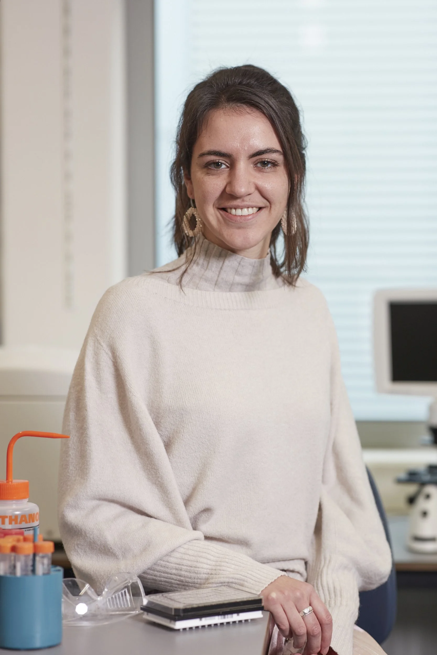 A woman in a cream sweater sitting at a desk in a laboratory with lab equipment and a computer in the background.