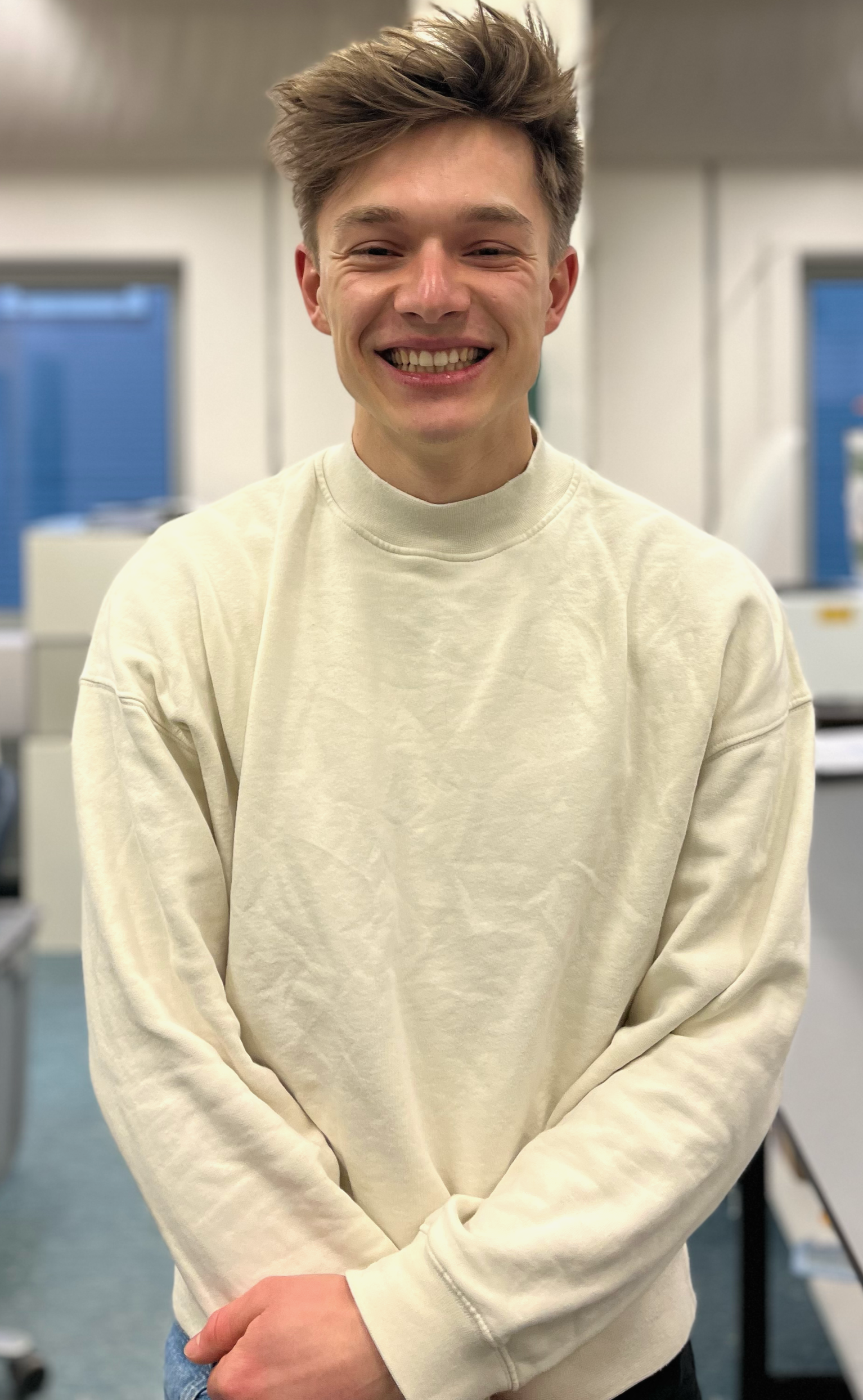 Smiling young man with tousled hair wearing a cream-colored sweatshirt, standing in an office setting.