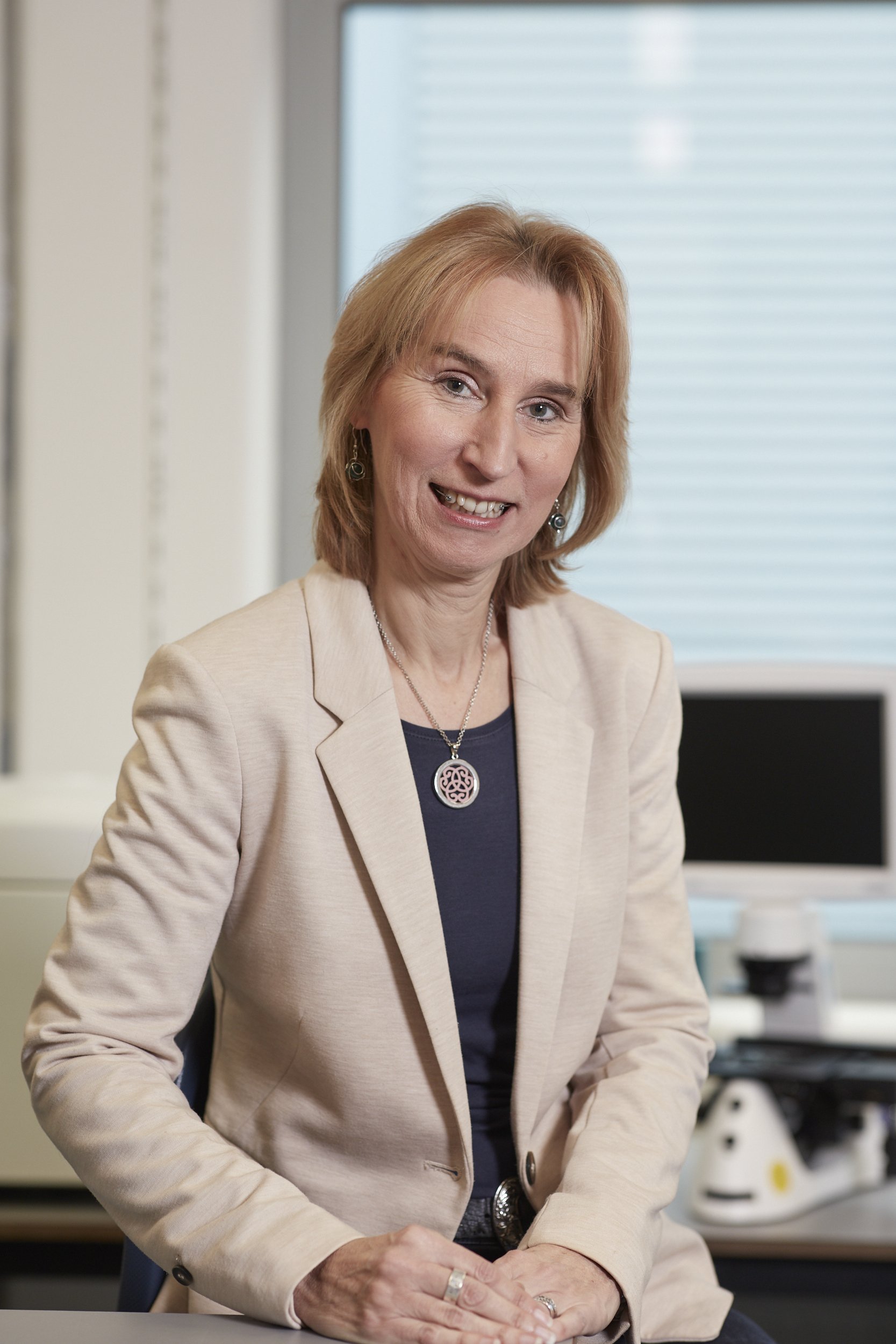 A middle-aged woman with shoulder-length red hair wearing a beige blazer, dark blue shirt, and jewelry, sitting at a desk in an office with a computer and window in the background.