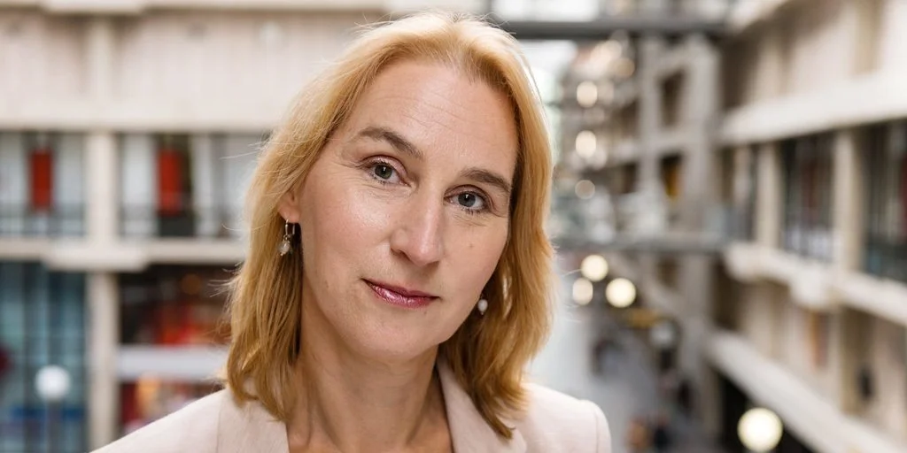 A woman with shoulder-length blonde hair, wearing earrings and a light-colored top, stands in front of a building with multiple balconies.