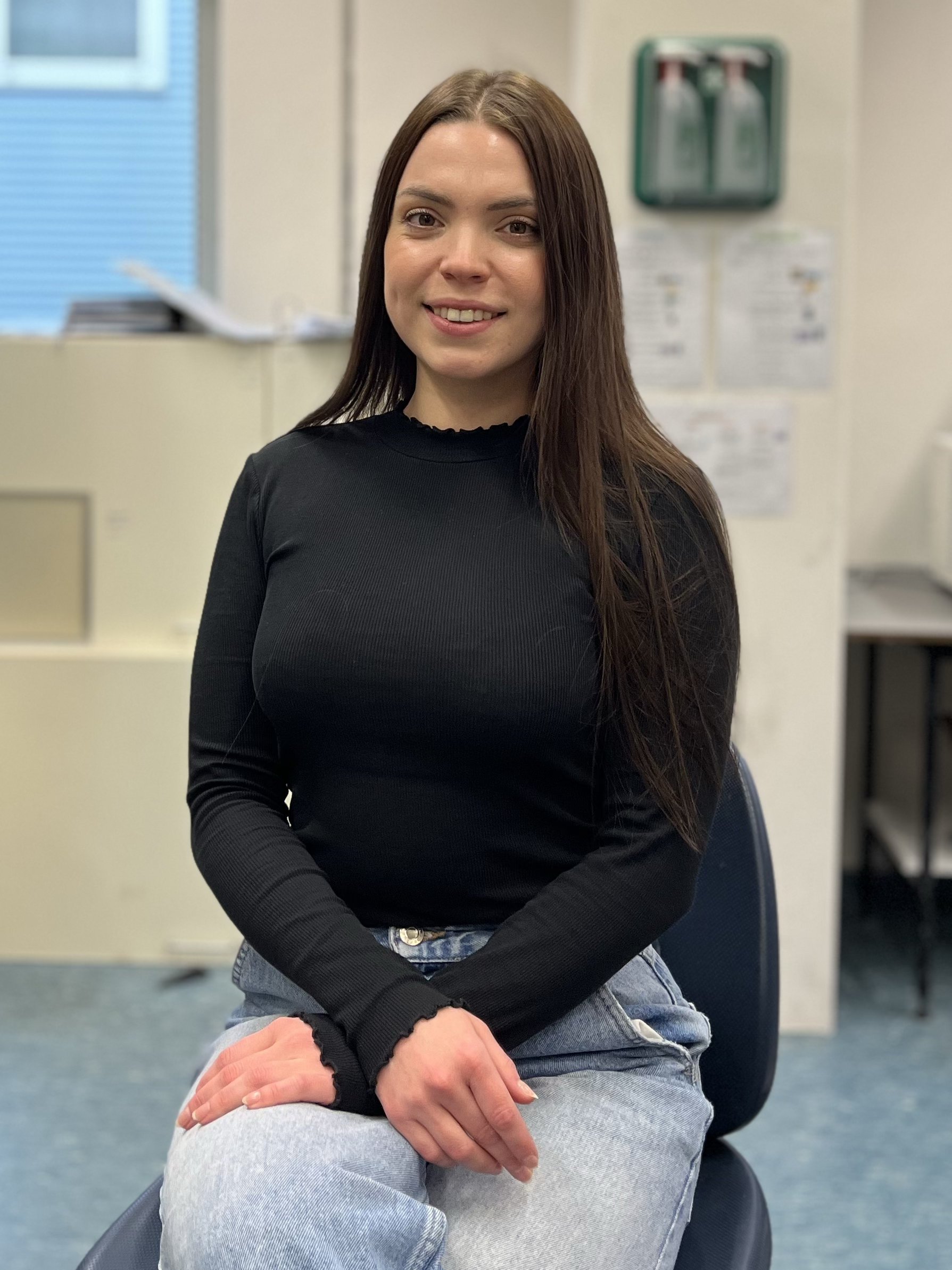 A young woman with long brown hair sitting on a chair in an office or classroom setting, smiling at the camera, wearing a black long sleeve shirt and light blue jeans.