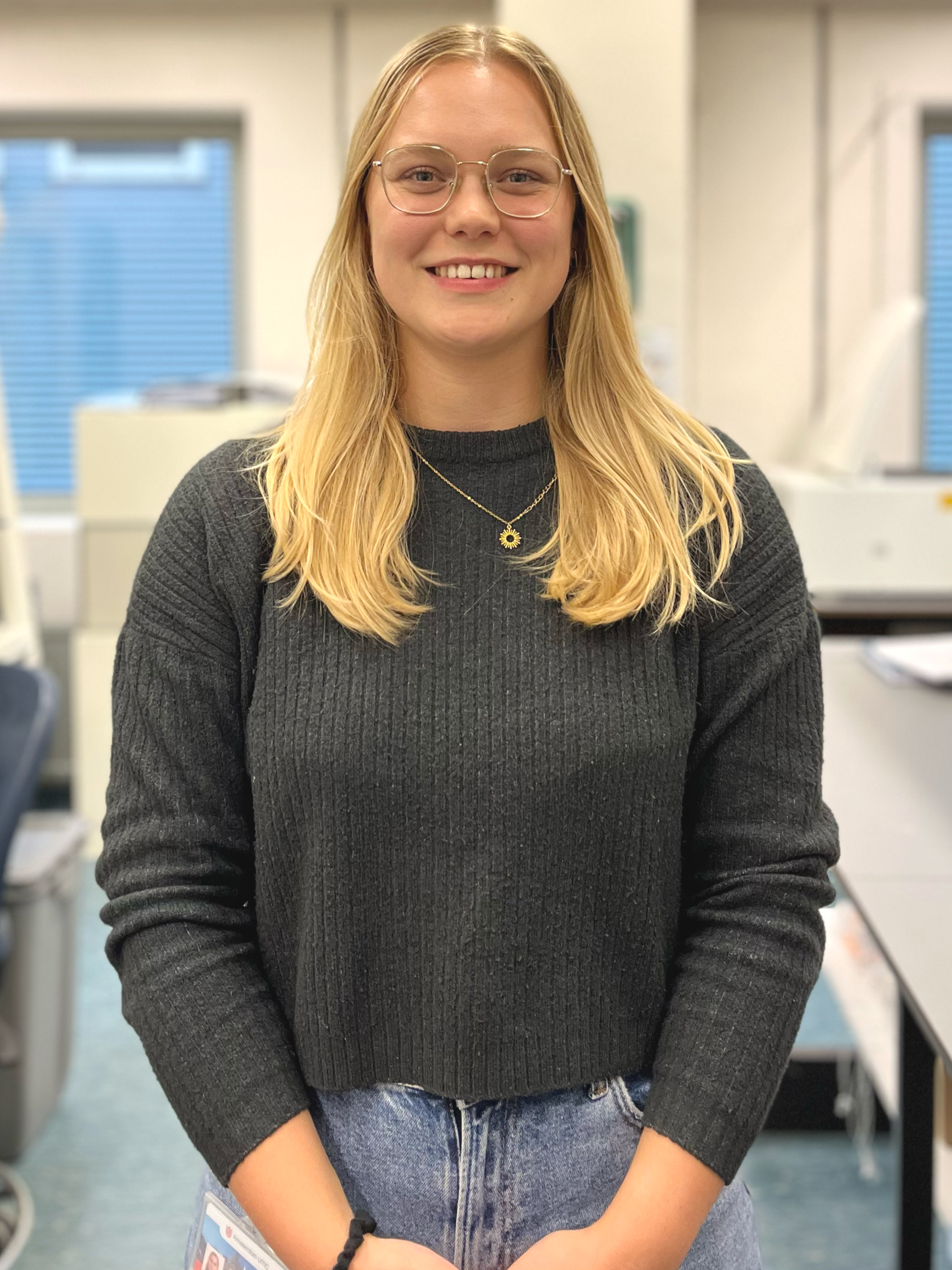 A young woman with long blonde hair, glasses, and a black ribbed sweater, smiling and standing in an office or laboratory setting.