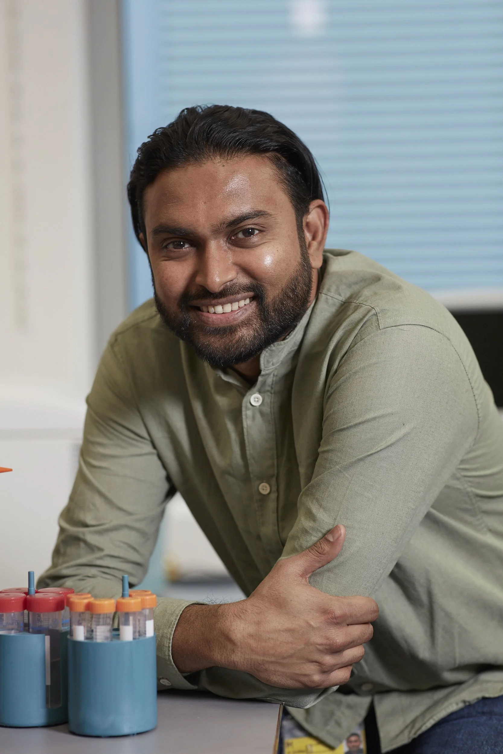 A smiling man with dark hair and beard leaning on a table with medical test tubes in blue holders, in a clinical setting.