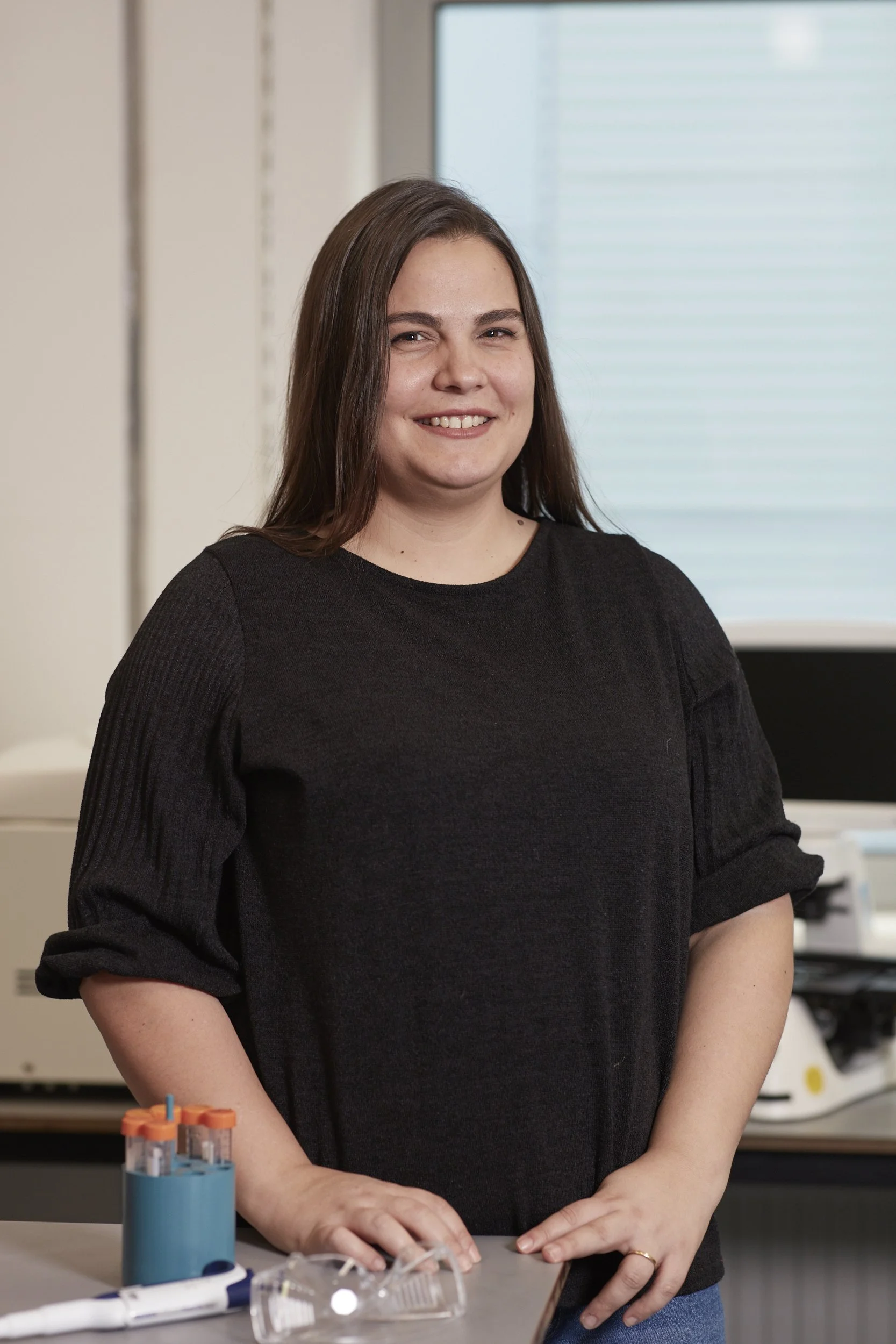 A smiling woman with long brown hair in a black top standing in a laboratory, with laboratory equipment on the counter in front of her.