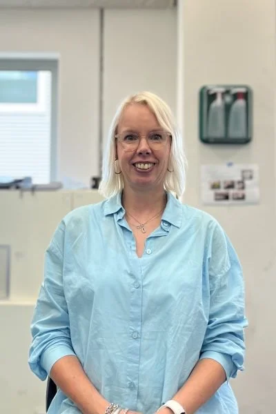 A woman with blonde hair, wearing glasses and a light blue shirt, is standing indoors and smiling at the camera.