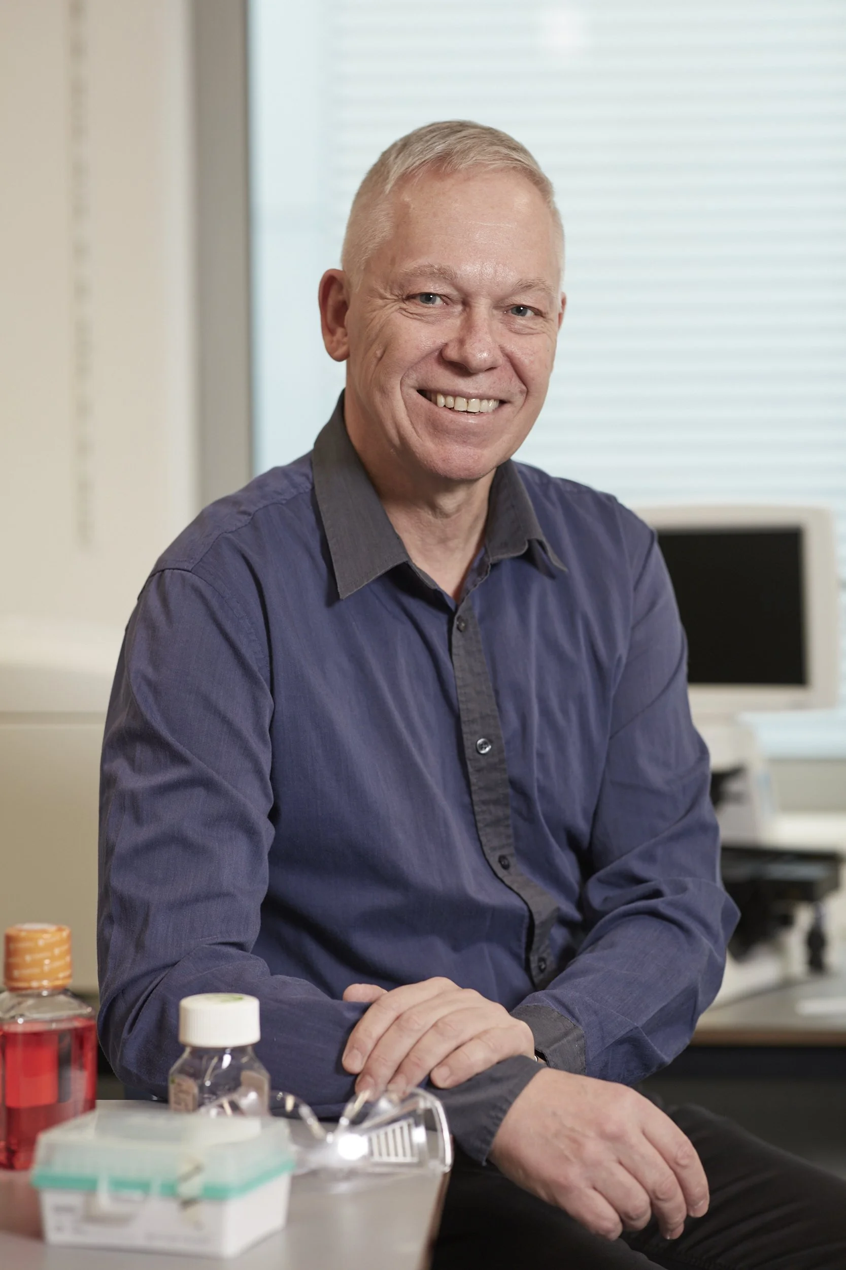 A smiling man with short gray hair, wearing a dark blue shirt, sitting at a desk with various medicines and pills in front of him, in an office setting.