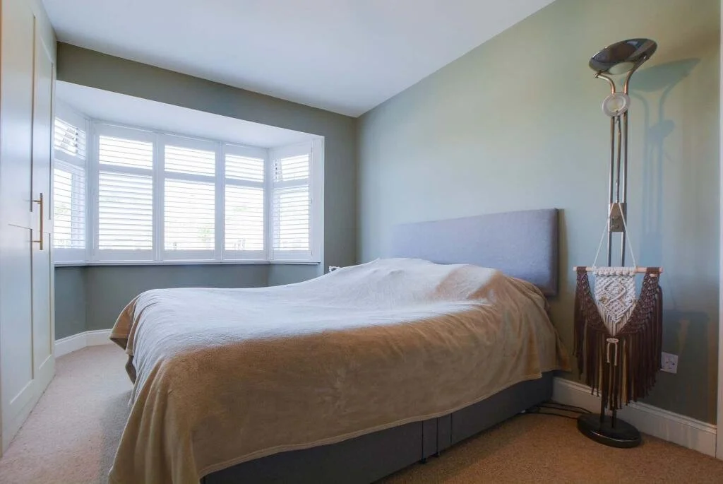 A bedroom with a bed covered in a beige blanket, grey headboard, large bay window with white shutters, and a tall floor lamp with a tapestry hanging from it.