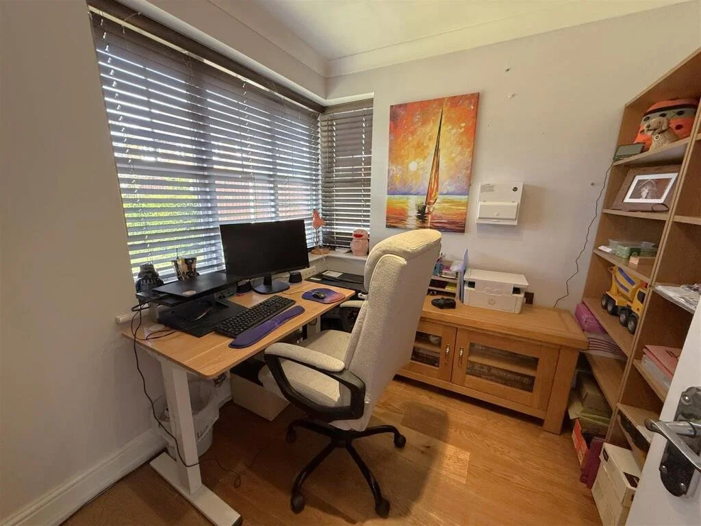 Home office with a desk, computer monitor, keyboard, mouse, and office chair, with large windows covered by blinds and a colorful sailboat painting on the wall.