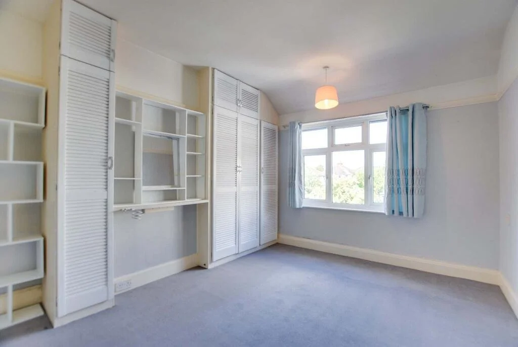 Empty bedroom with built-in white shelves and closet doors, a window with blue curtains, and beige walls and carpet.