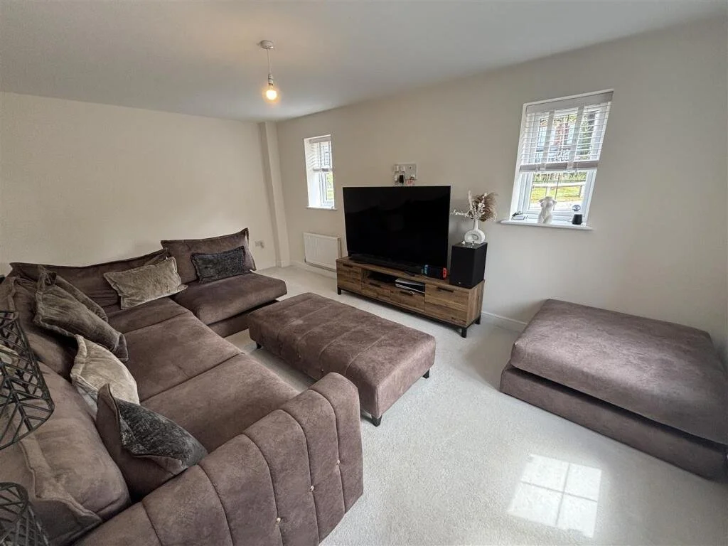 Living room with a large brown sectional sofa, a matching ottoman, a wooden TV stand with a large flat-screen TV, two small side tables with decorative items, and two windows with white blinds, all on white walls and carpeted floor.