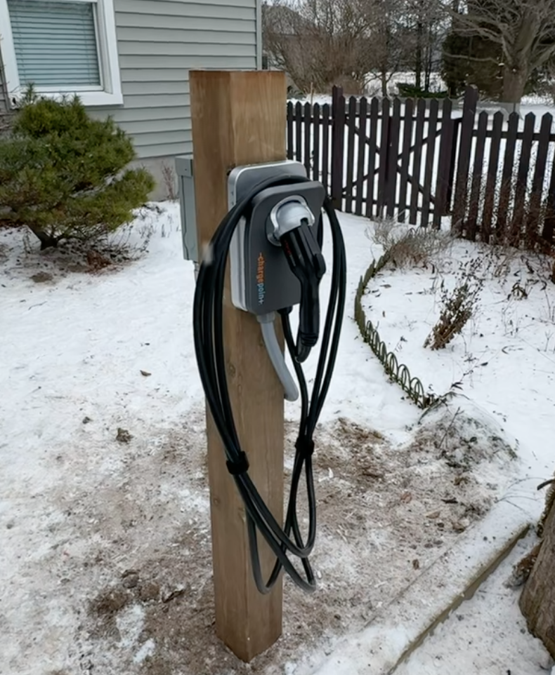 Electric vehicle charging station mounted on a wooden post outdoors in a snowy yard.
