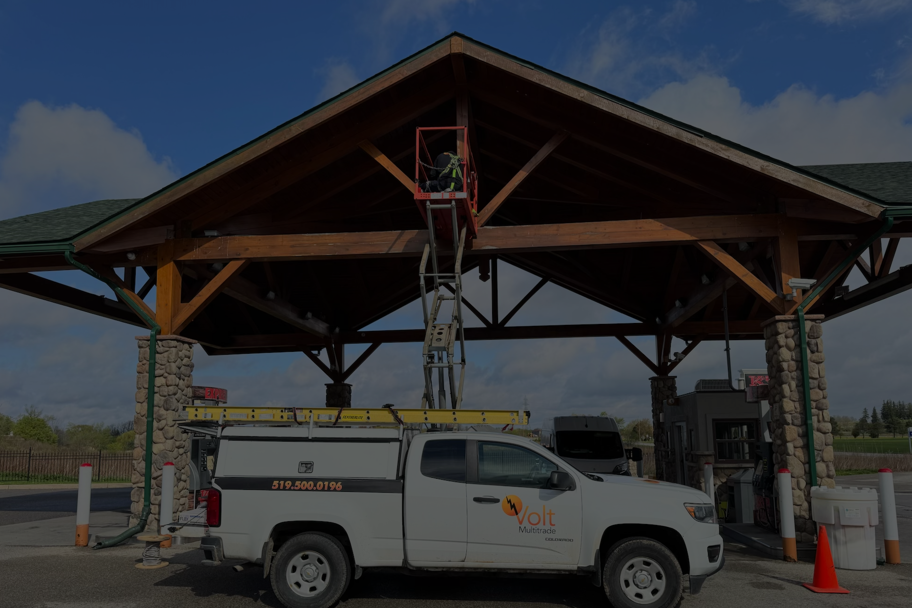 A worker on a raised platform working near the peak of a wooden gazebo structure, with a service vehicle parked underneath.