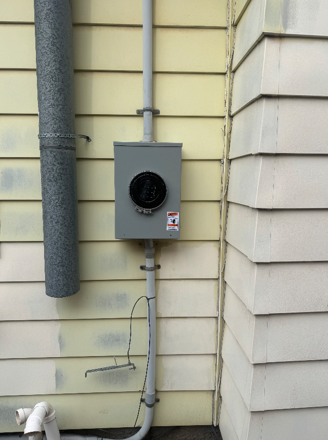 Electrical meter box mounted on the wall of a house, with gray siding and a black pipe on the left.