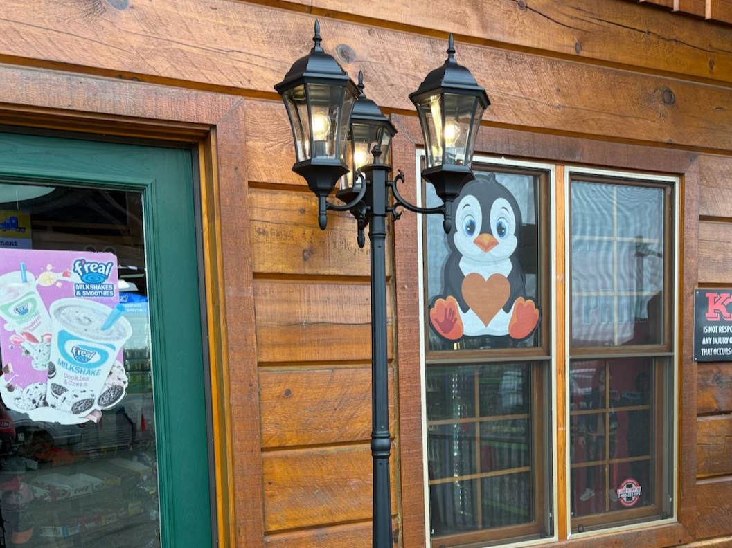 A wooden building with green and glass door, two large windows, a decorative black lamp post with four lanterns, and a penguin cartoon sticker on the window showing a penguin holding a heart.