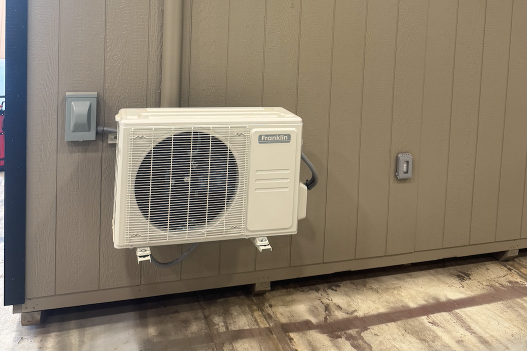 Exterior wall with a white Franklin air conditioning or heat pump unit mounted on it, next to a gray electrical outlet and an electrical switch.