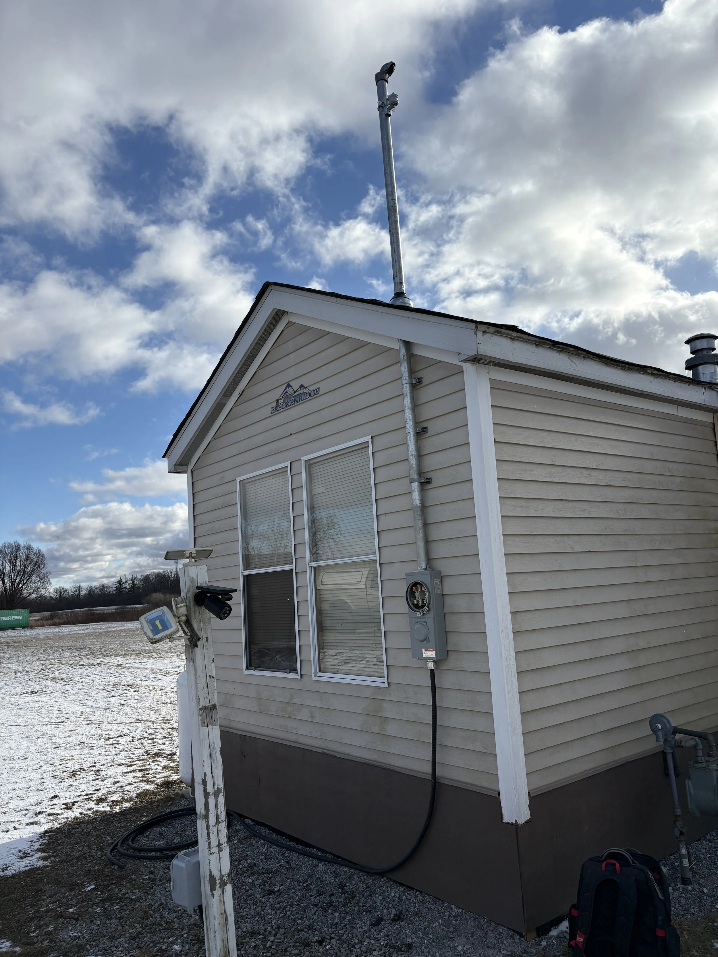 A small house with beige siding, two windows, and an electrical meter on the side. A metal pipe extends from the roof, and there is a weather station with a white pole and camera at the front. The sky is partly cloudy with blue sky visible.