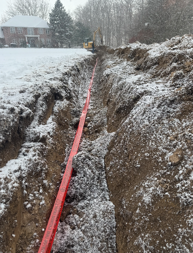 A construction site in a snowy area with a large excavation trench. A red pipe runs through the trench, and a yellow excavator is visible at the far end. There are snow-covered trees and a house in the background.