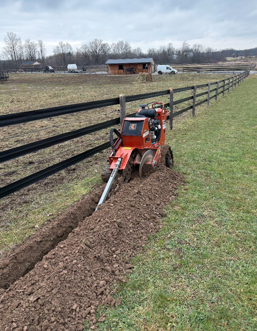 A red trenching machine digging a trench along a wooden fence on a farm or ranch, with a barn, hay bales, and vehicles in the background on a cloudy day.