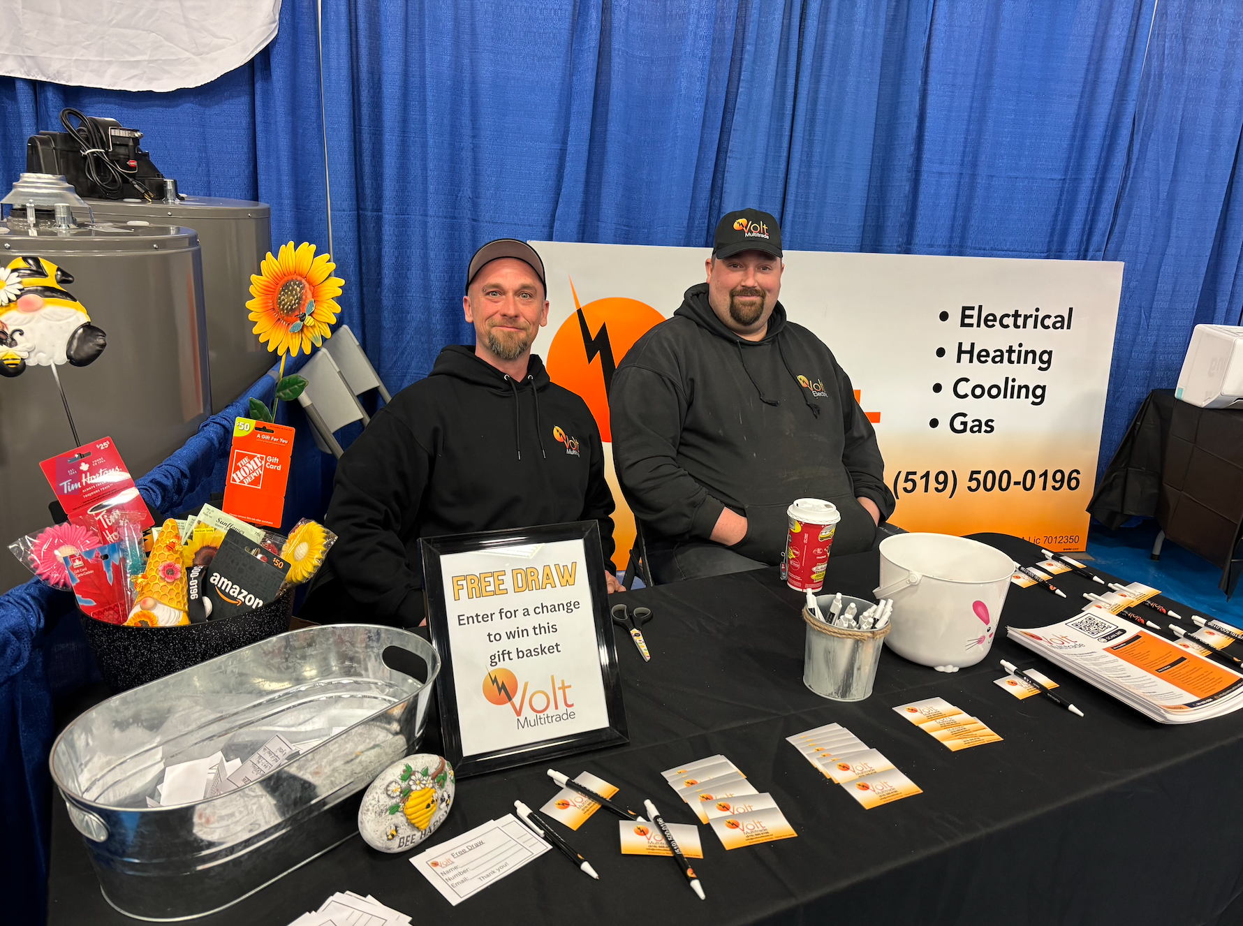 Two men sitting behind a table at a promotional booth, with a large sign displaying electrical services, and a sign announcing a free raffle drawing. The table has pens, flyers, and a small donation box, with gift cards and small items nearby.