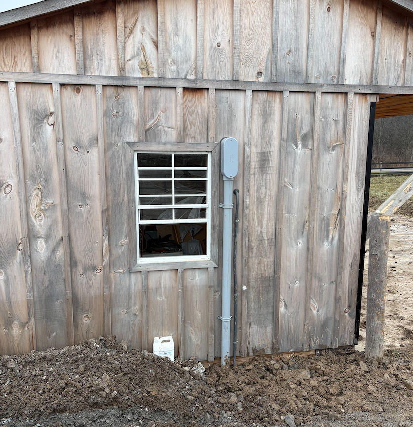 Close-up of a wooden exterior wall of a building with a small window that has metal bars. There is electrical conduit and a gray electrical box mounted on the wall. The ground is disturbed dirt with a white plastic container nearby.