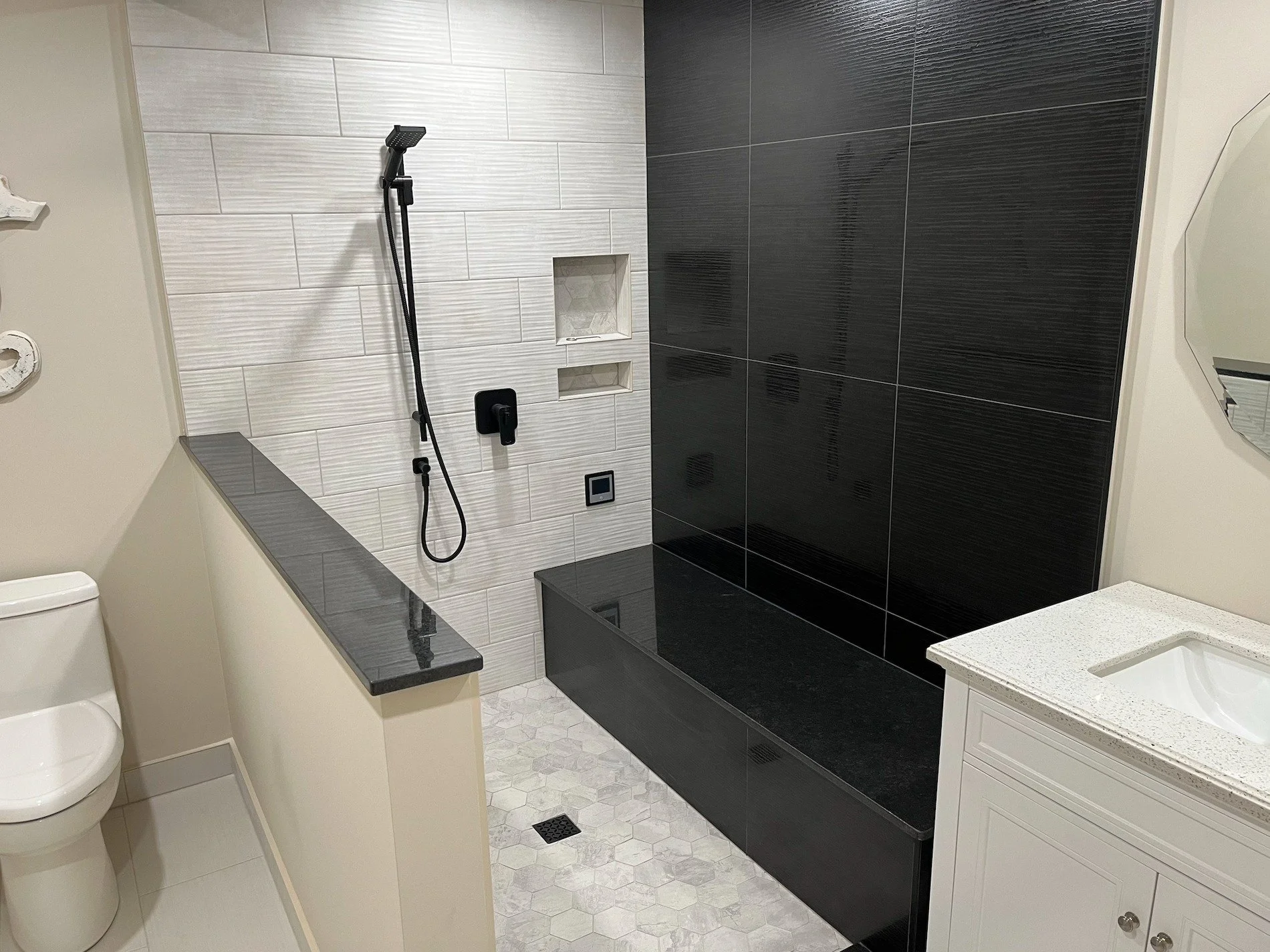 Modern bathroom with a shower area featuring a black bench and black wall panels, a partial view of a toilet and a white vanity with a gray speckled countertop and an undermount sink.