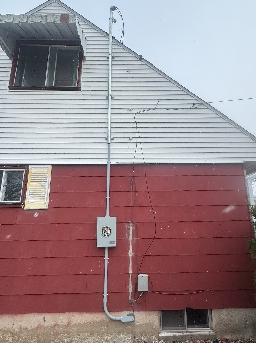 Exterior of a two-story house with white upper siding and red lower siding. Visible electrical meter box, conduit pipe, and wires attached to the house. A small basement window at the bottom.