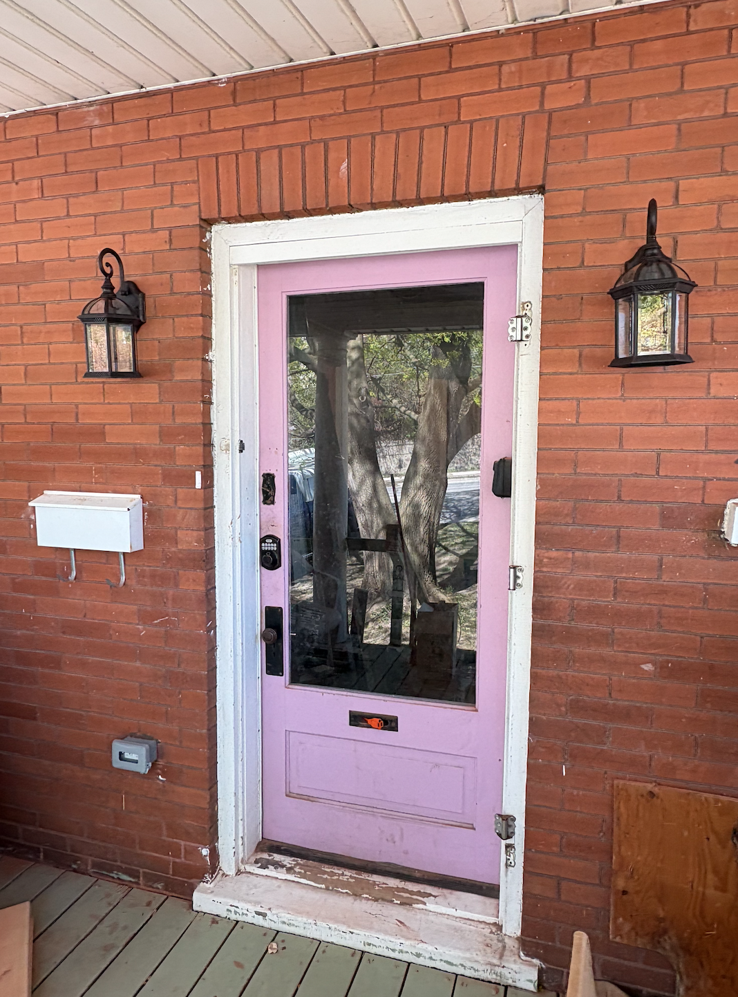 A pink front door with a glass panel, installed on a red brick house, flanked by two black lantern-style exterior lights. A white mailbox is mounted on the brick wall to the left of the door. The door has electronic lock and keypad, and a smaller mai