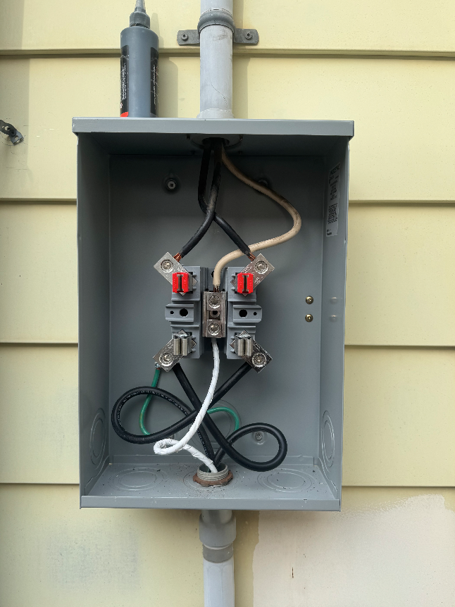 Electrical meter box with wiring and circuit breakers inside, mounted on a house exterior wall.