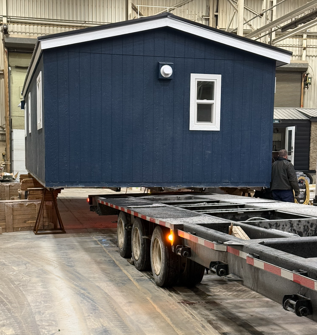 A blue tiny house being transported on a flatbed trailer inside a warehouse. The house has white trim around the door and window and a pipe on the side. A person in a black jacket is standing near the house.