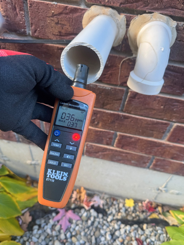 Person wearing a glove using a carbon monoxide meter to measure gas levels inside a white pipe on a brick wall outside.