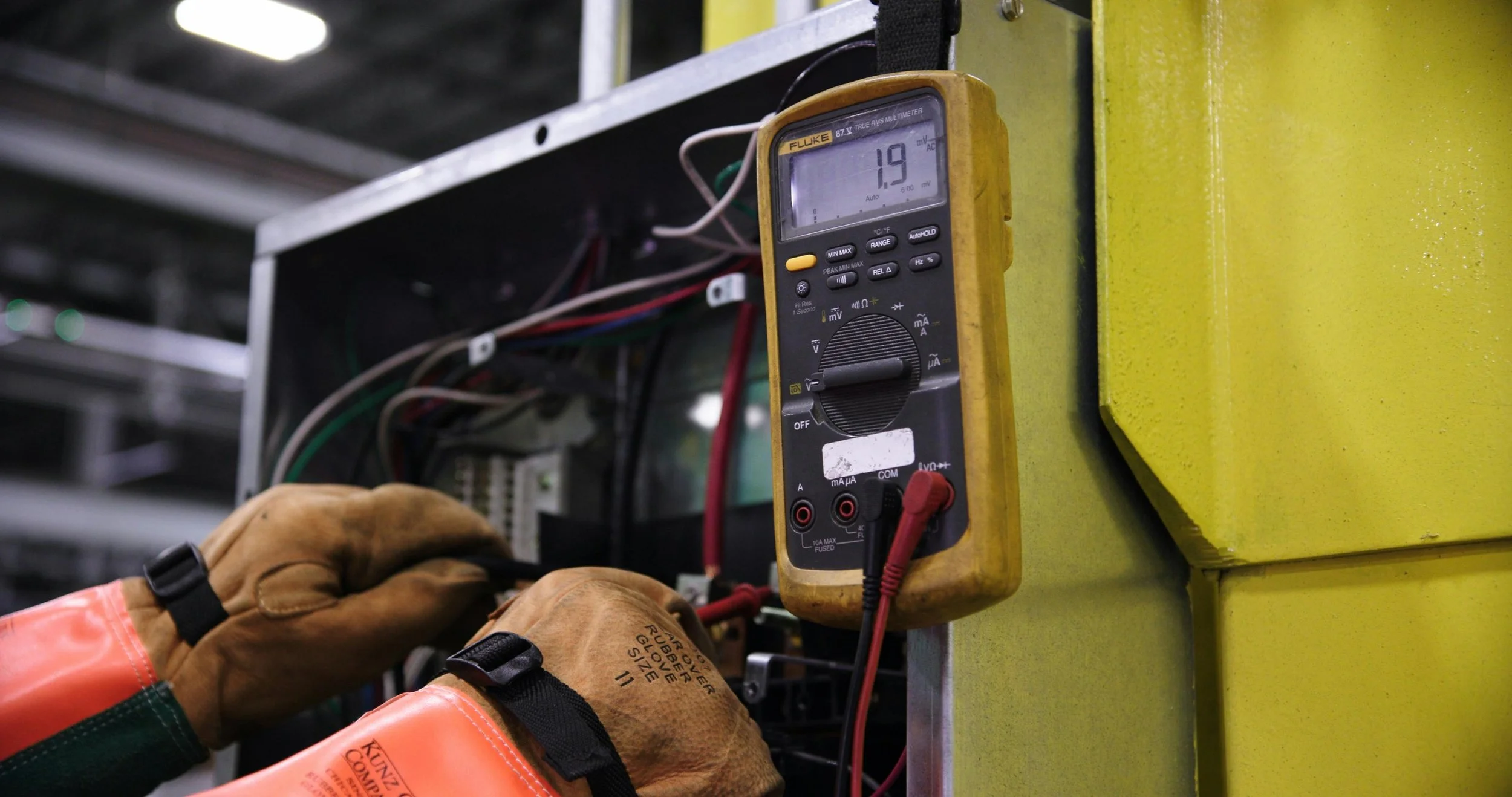 A person wearing gloves using a multimeter to measure electrical voltage or current in an industrial electrical panel.