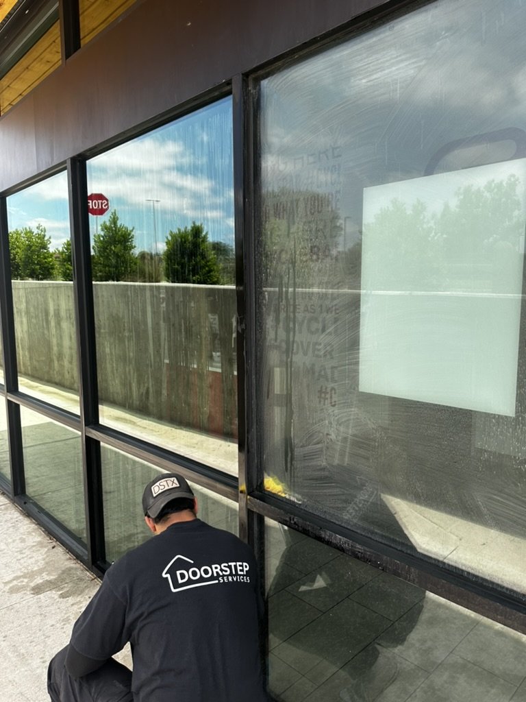 A repair technician kneeling outside a glass-front store, wearing a black shirt and cap with the Doorstep Services logo, working on the lower part of a window frame.