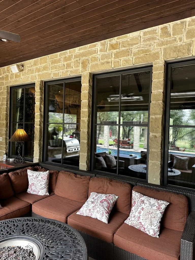 Outdoor patio area with a brown wicker sectional sofa with floral pillows, a fire pit table, and large windows showing a backyard with a pool and patio furniture.