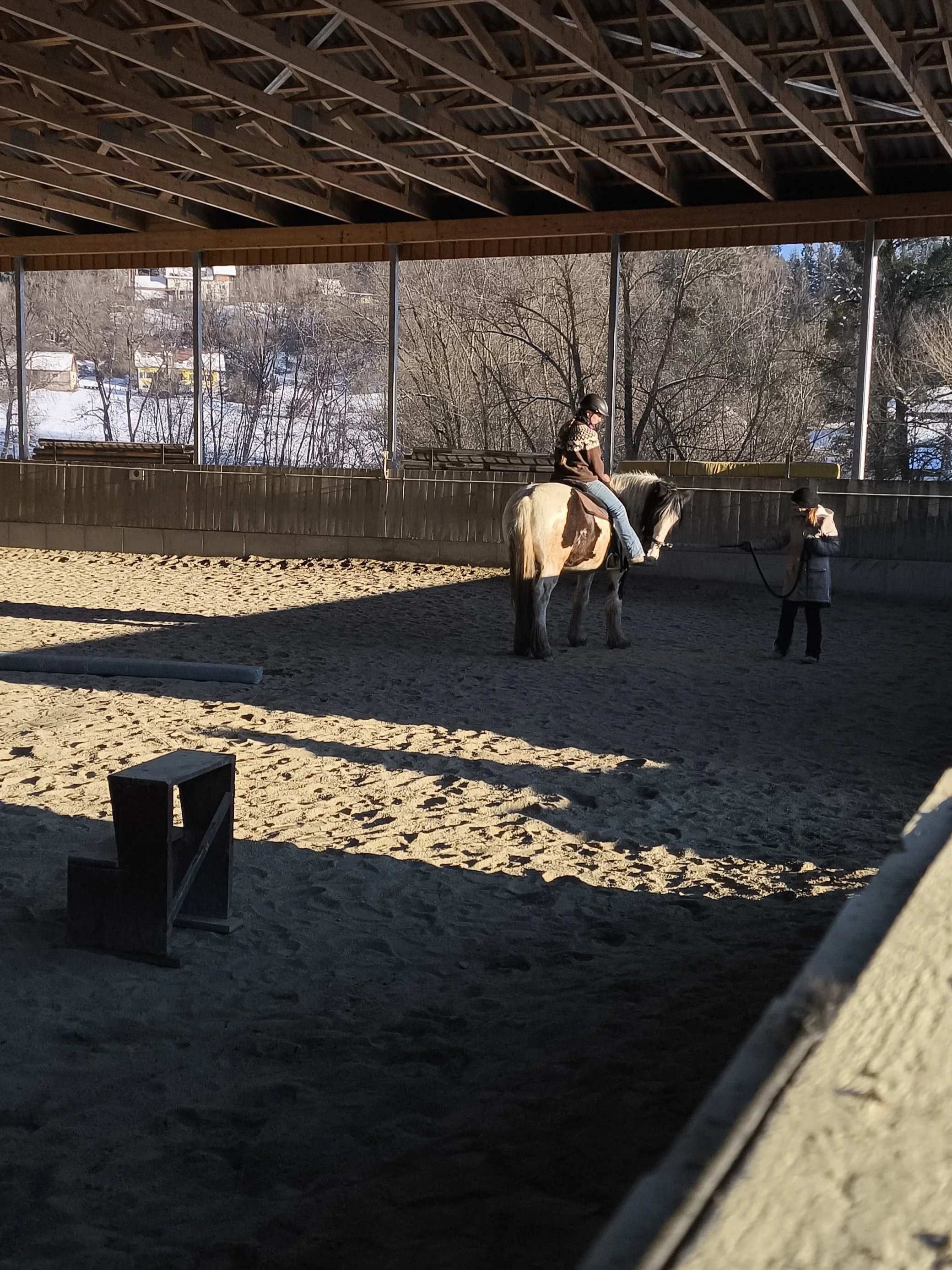 Ein Mädchen reitet auf einem weißen und braunen Pferd in einer Reithalle, während eine Frau das Tier führt. Im Hintergrund sind Bäume und verschneite Häuser sichtbar.