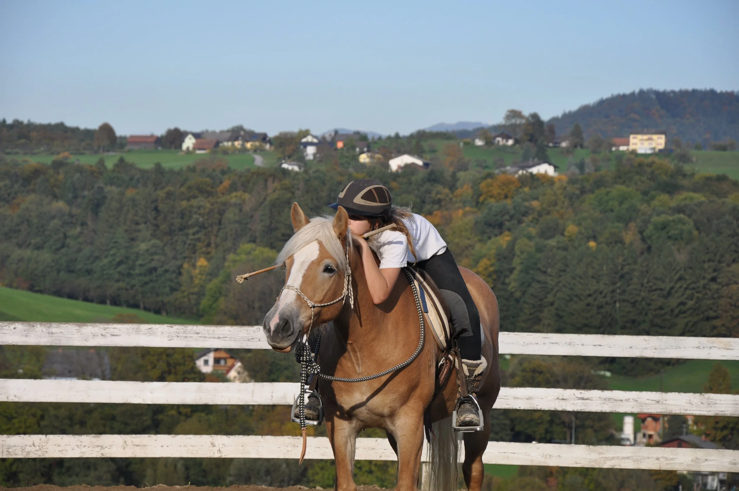 Ein Mädchen reitet auf einem Pferd, umarmt es liebevoll und zeigt Zuneigung auf einer Reitausbildung im Freien mit einer ländlichen Landschaft im Hintergrund.