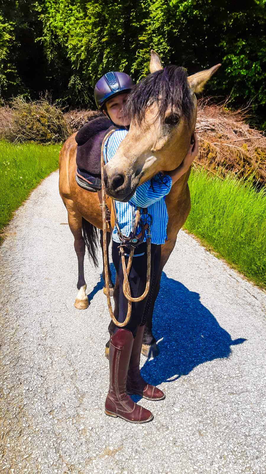 Ein Kind mit Reithelm, umarmt ein Pferd auf einem Weg in einer grünen, bewaldeten Umgebung.