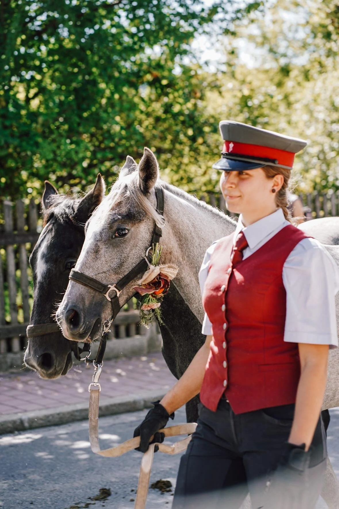 Eine junge Frau in Uniform mit einem roten Reitweste und Mütze reitet und führt zwei Pferde, die mit Blumen geschmückt sind, in einer grünen, sonnigen Umgebung.