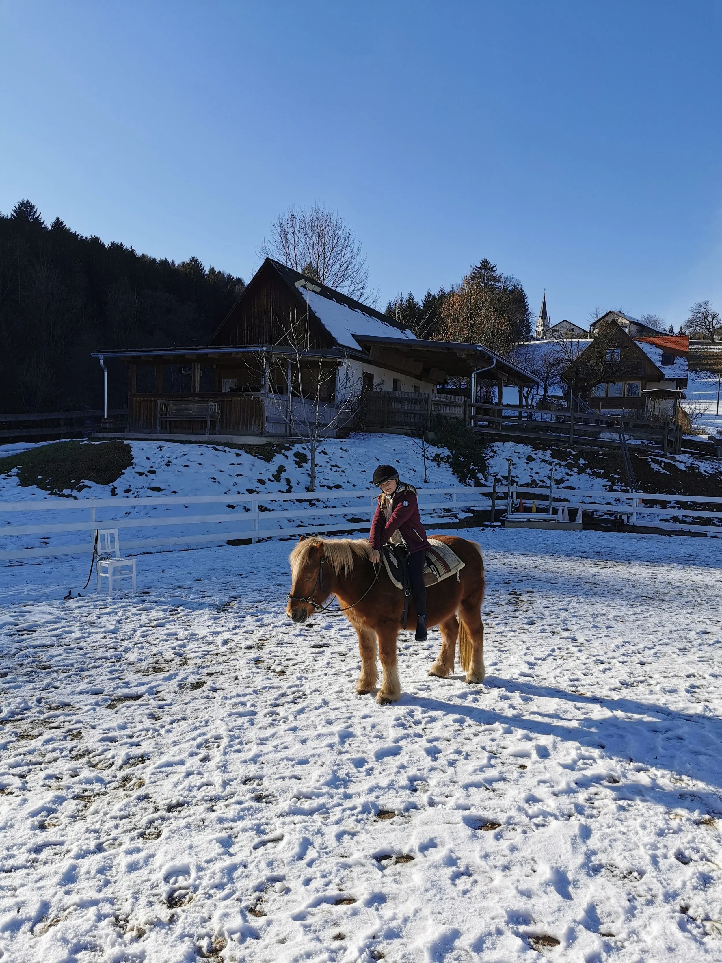 Ein Kind reitet auf einem Pony im Schnee vor einem Haus im ländlichen Gebiet bei sonnigem Wetter.
