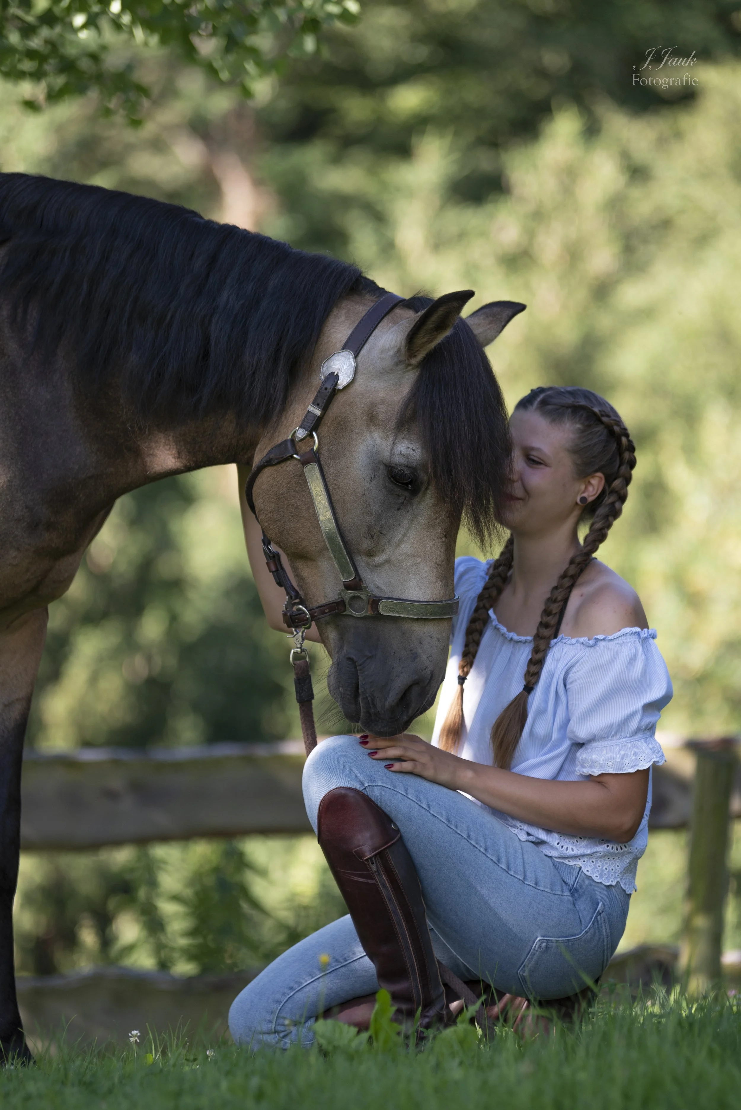 Junge Frau in Bluejeans und braunen Stiefeln sitzt vor einem Zaun. Daneben ein Sandbraunes Pferd mit dunklem Maul. Es ist draußen in der Natur mit grünen Bäumen im Hintergrund.