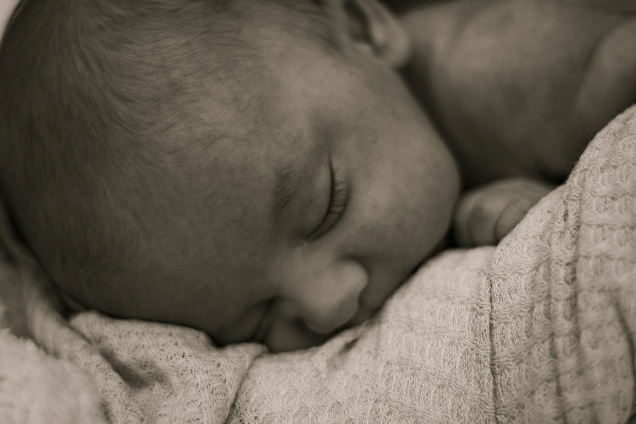 A close-up black and white photo of a sleeping newborn baby resting on a textured blanket.