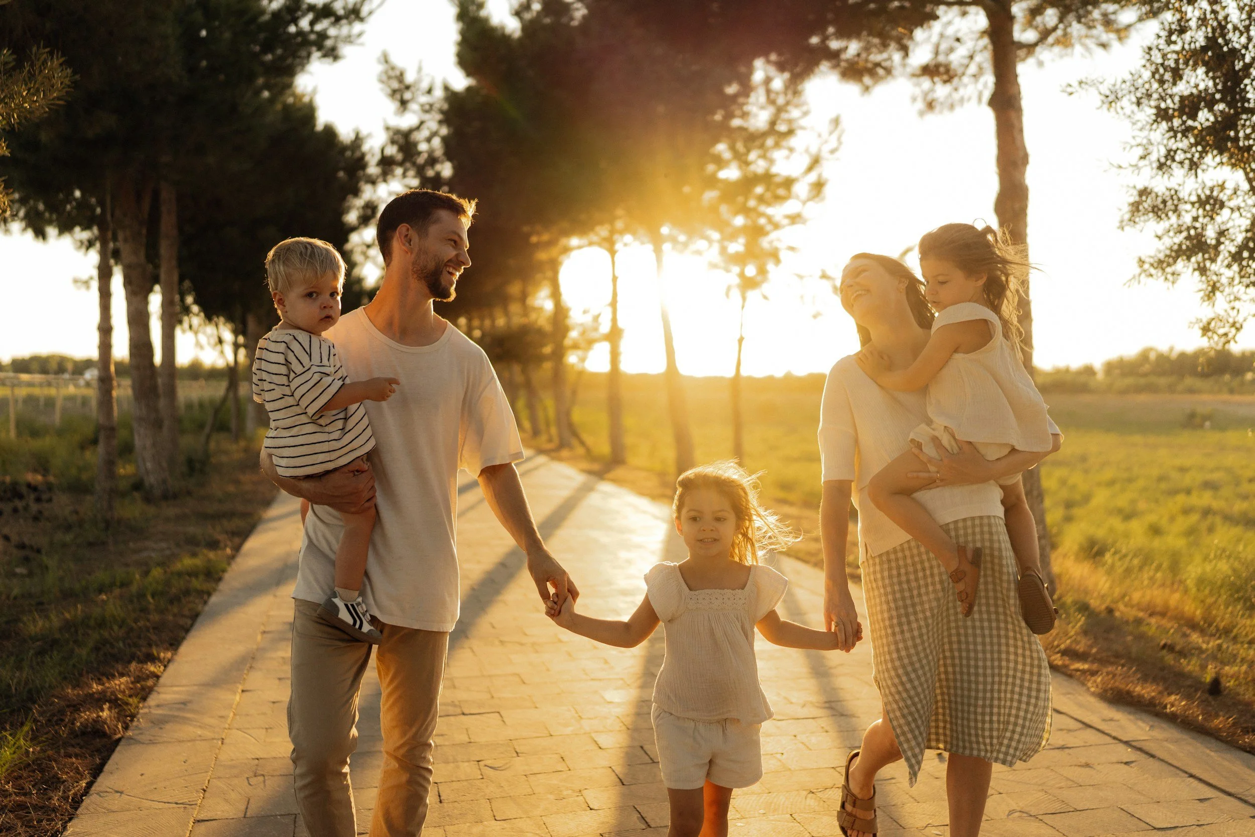 A happy family walking on a path in a park during sunset, with two children and two adults smiling and holding hands.