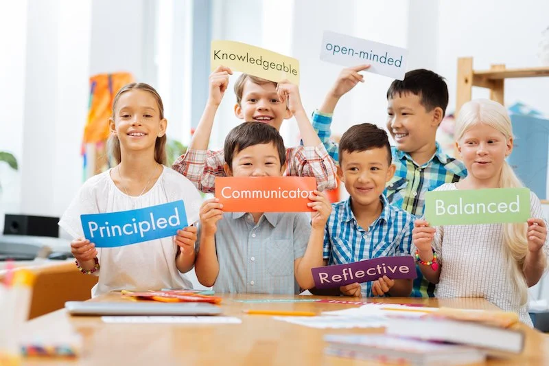 Group of six children in a classroom holding colorful signs with words like 'Principled,' 'Knowledgeable,' 'Open-minded,' 'Communicative,' 'Balanced,' and 'Reflective'.