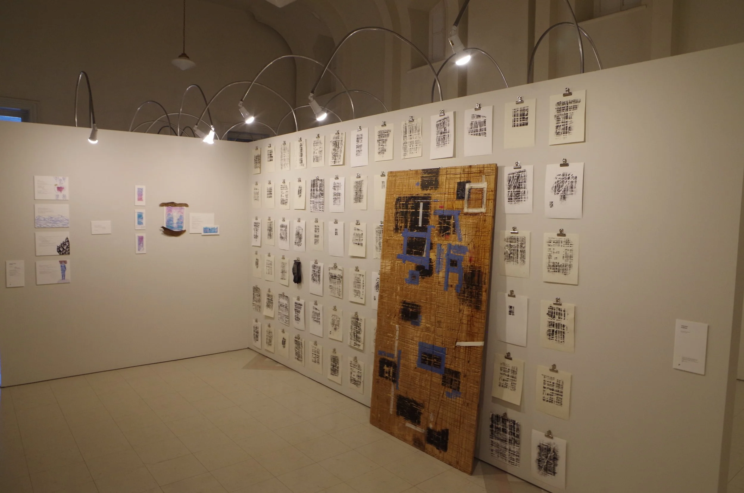 Exposition d'art avec plusieurs œuvres et documents affichés sur un mur blanc, incluant un tableau en bois avec des éléments bleu et noir, éclairage dirigé par des lampes modernes.