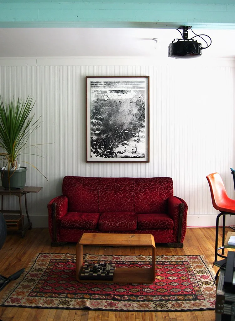 Salon avec un canapé rouge en velours, une table en bois, un tapis aux motifs géométriques, un tableau noir et blanc au mur, une plante verte à gauche, et un projecteur suspendu au plafond.