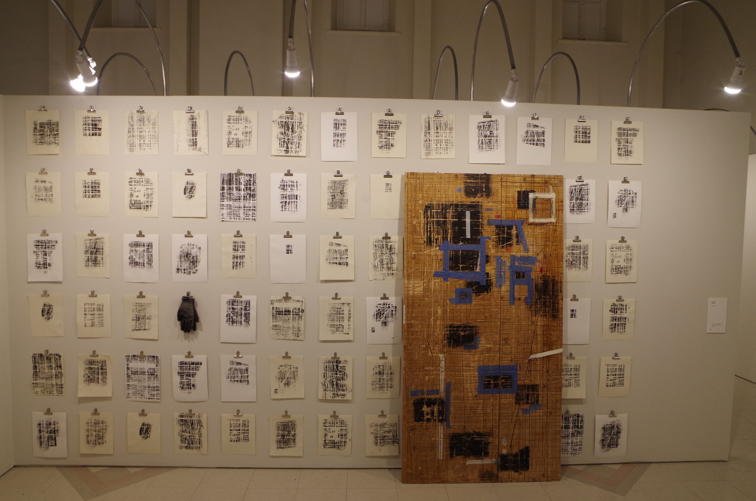 Une exposition d'art avec plusieurs feuilles imprimées en noir et blanc accrochées à un mur, accompagnée d'une peinture en bois avec des détails géométriques bleus, encadrée par des lampes suspendues.