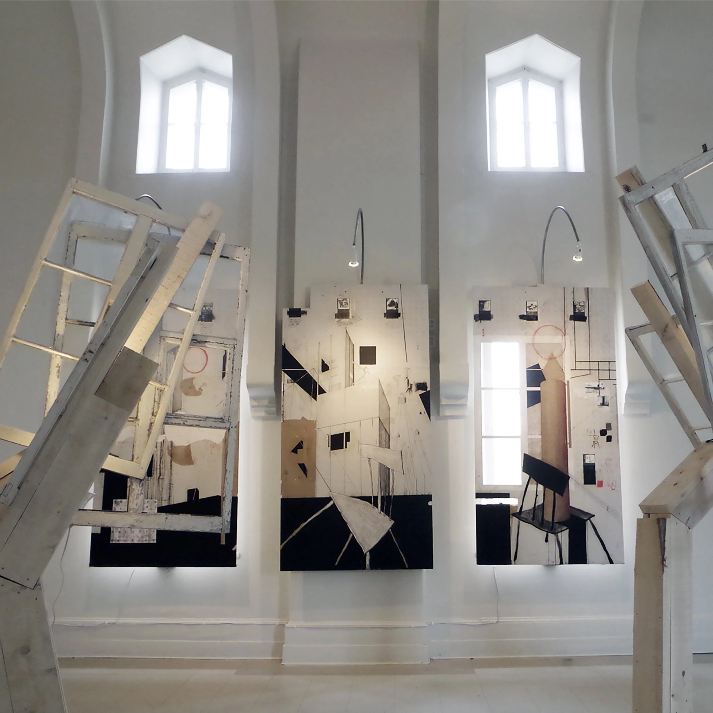 Trois œuvres d'art abstrait suspendues au mur d'une galerie, entourées de structures en bois inclinées et éclairées par des lampes, avec des fenêtres en haut laissant entrer la lumière