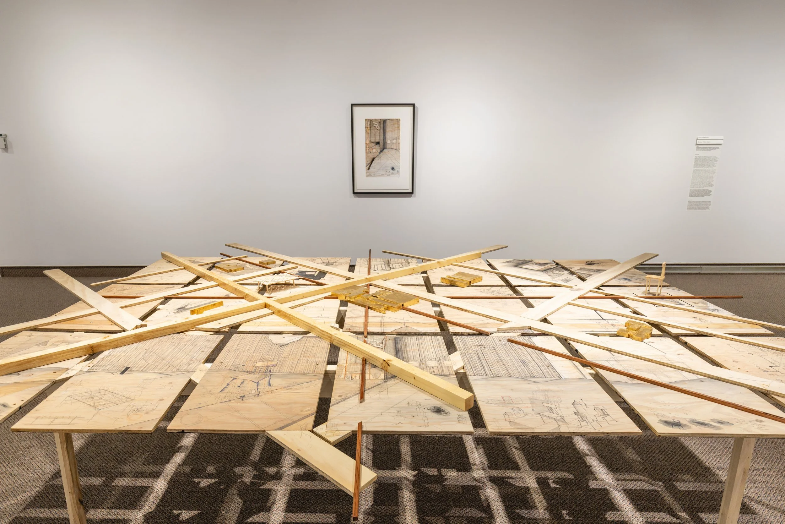 Une œuvre d'art composée de sketches, plans et pièces en bois disposés sur une table dans un musée, avec un tableau encadré sur le mur derrière.