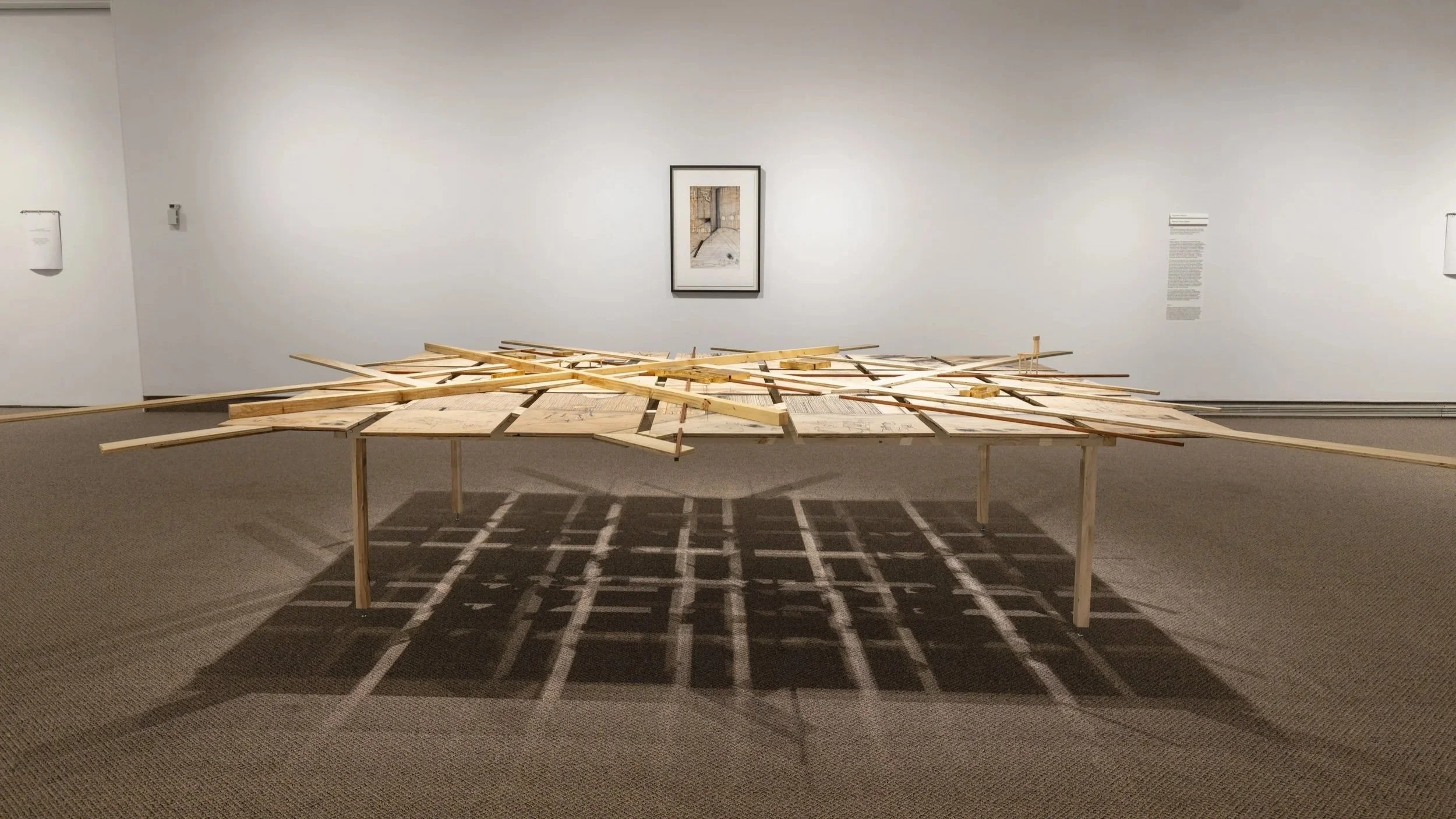 Installation artistique en bois , dans une galerie d'art, avec une photo encadrée au mur en arrière-plan.