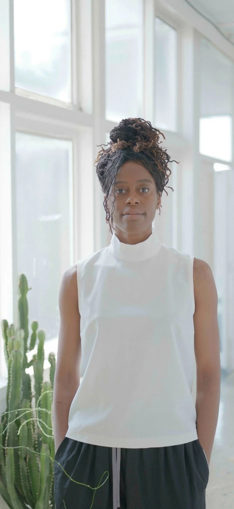 A woman with natural curly hair in a high bun, wearing a sleeveless white top and black pants, standing in a bright room with large windows and a cactus in front of her.
