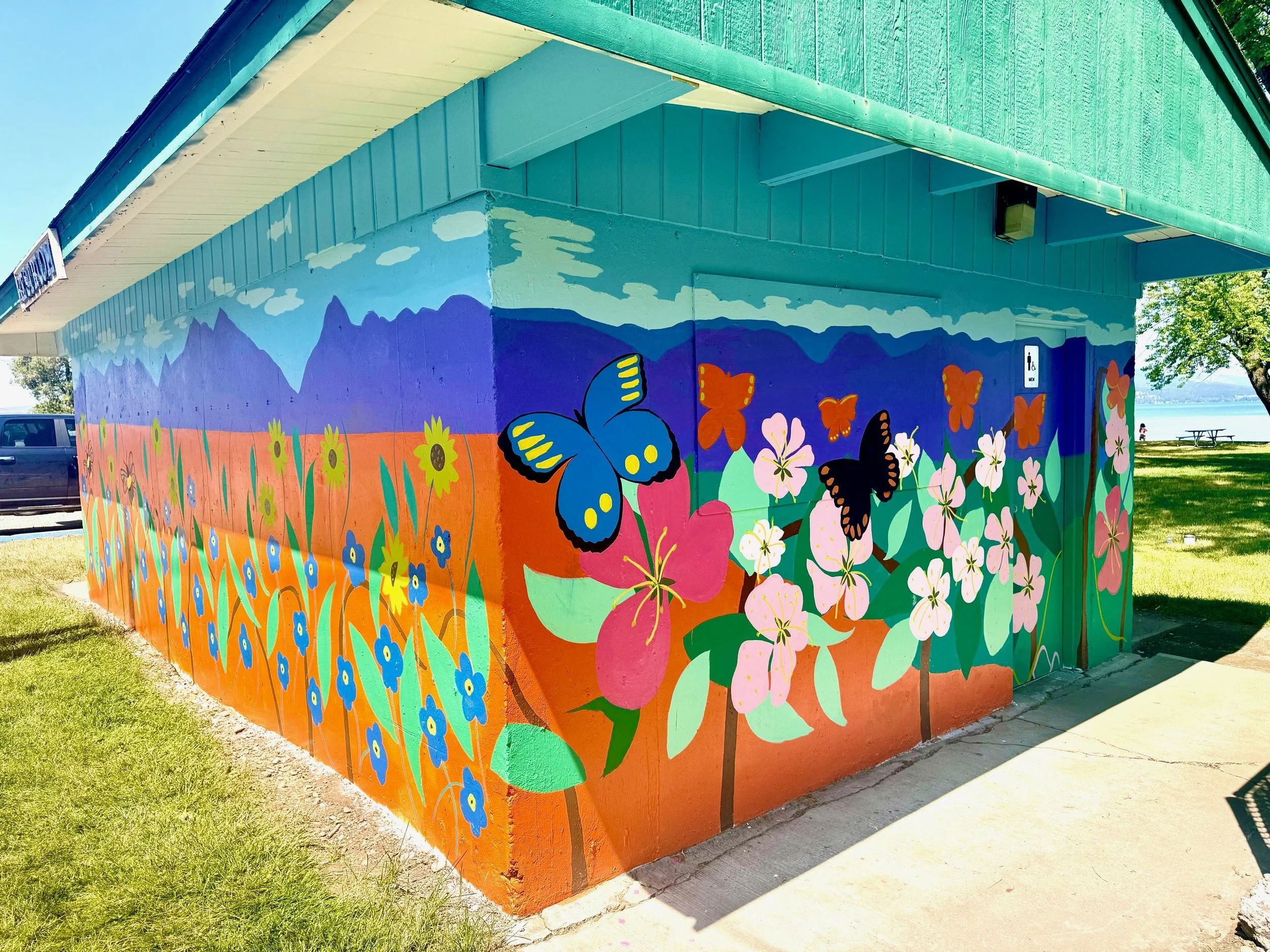 Colorful mural of flowers and butterflies on the side of a building near a park with a lake in the background.